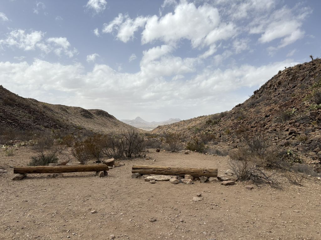 A desert valley with rocky hills and two wooden log benches in the foreground under a cloudy sky.